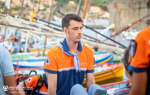 Un secouriste bénévole est concentré en brancardant une victime, en fond on aperçoit des barques catalanes et la ville de Collioure
